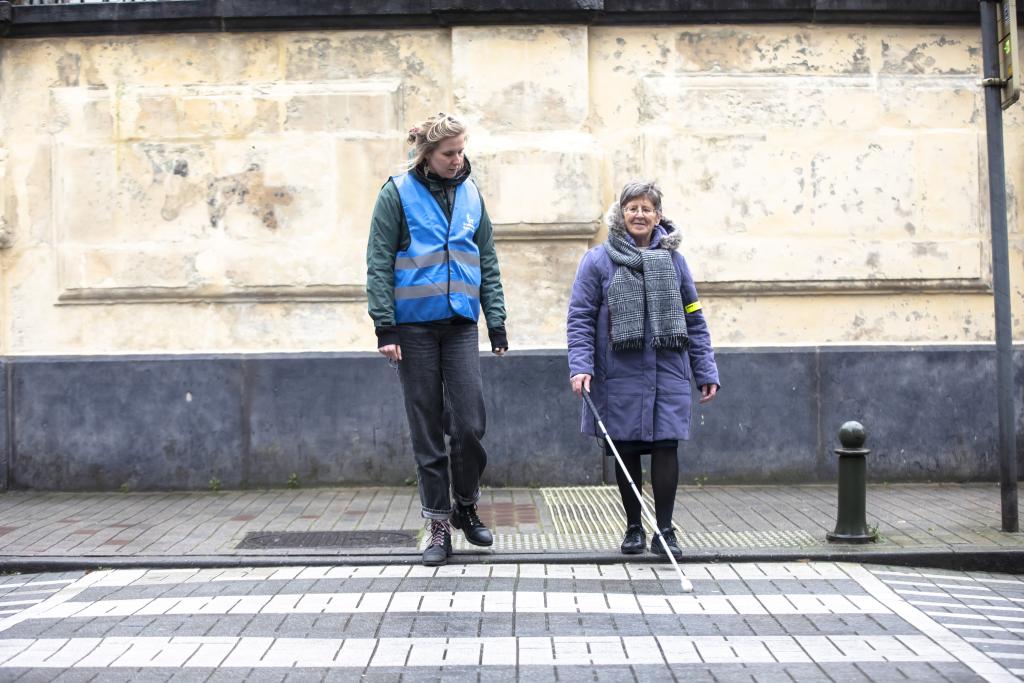 Photo d'une dame qui se prépare à traverser sur le passage clouté, à ses côtés, une thérapeute de la Ligue Braille la guide.